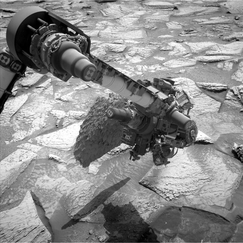 Robotic arm of a Mars rover hovers over cracked, layered rocks on the Martian surface, with the arm’s instruments extended and shadow visible among the textured terrain.