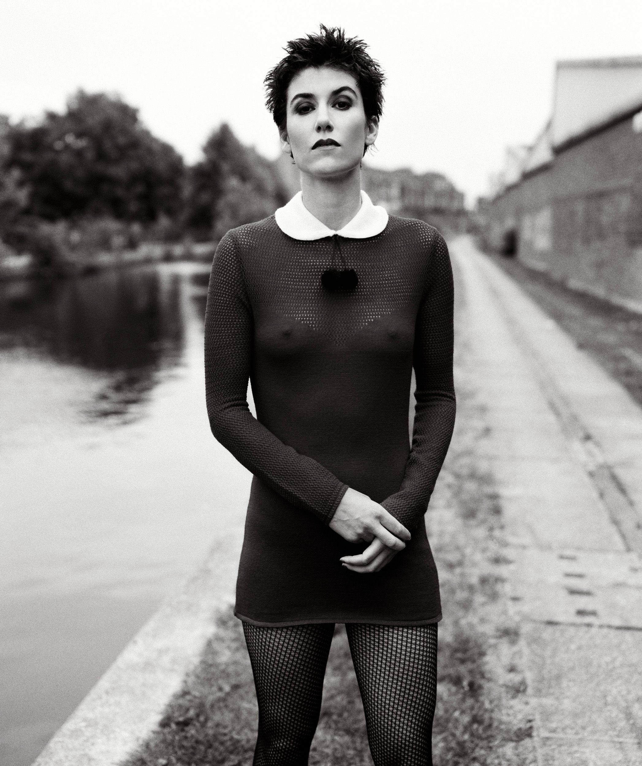 A black and white full-body shot of a woman with short, dark hair standing by a canal, wearing a dark, long-sleeved mini dress with a white collar and patterned tights.
