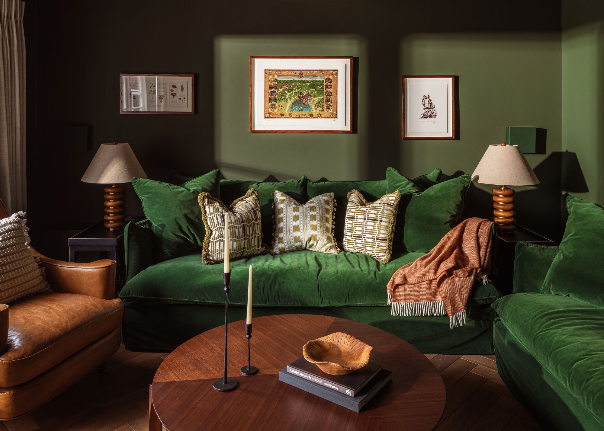 A cozy snug room with dark moss green walls, a dark bottle green velvet corner sofa with patterned cushions, and a red throw. A round wooden coffee table, two lamps, and three artworks on the wall.