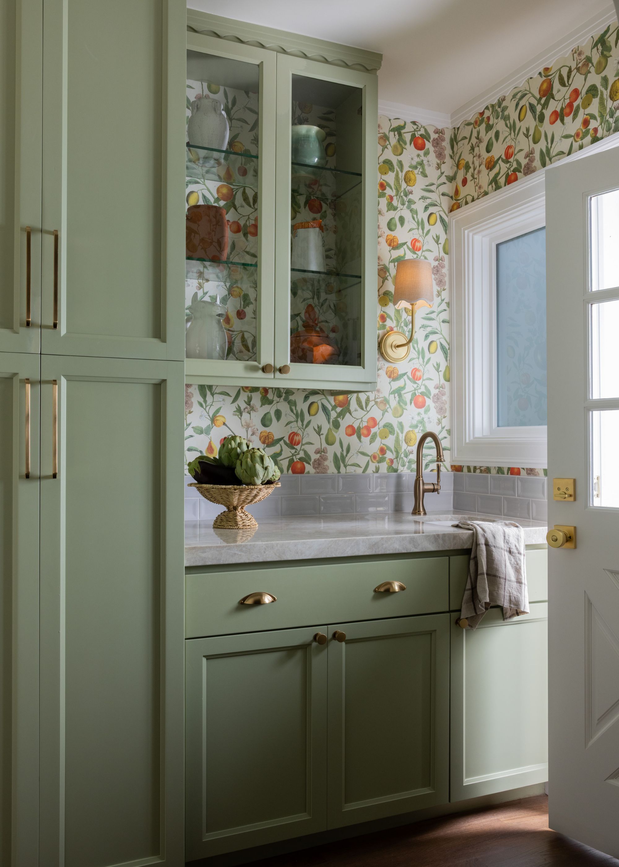 A small kitchen with sage green cabinets, gray countertops, and a green and orange botanical motif wallpaper on the walls.