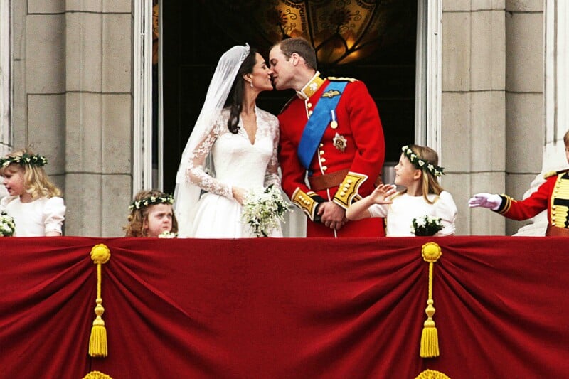 A bride and groom in formal wedding attire kiss on a balcony draped with red cloth and gold tassels, surrounded by young flower girls in white dresses and floral crowns.
