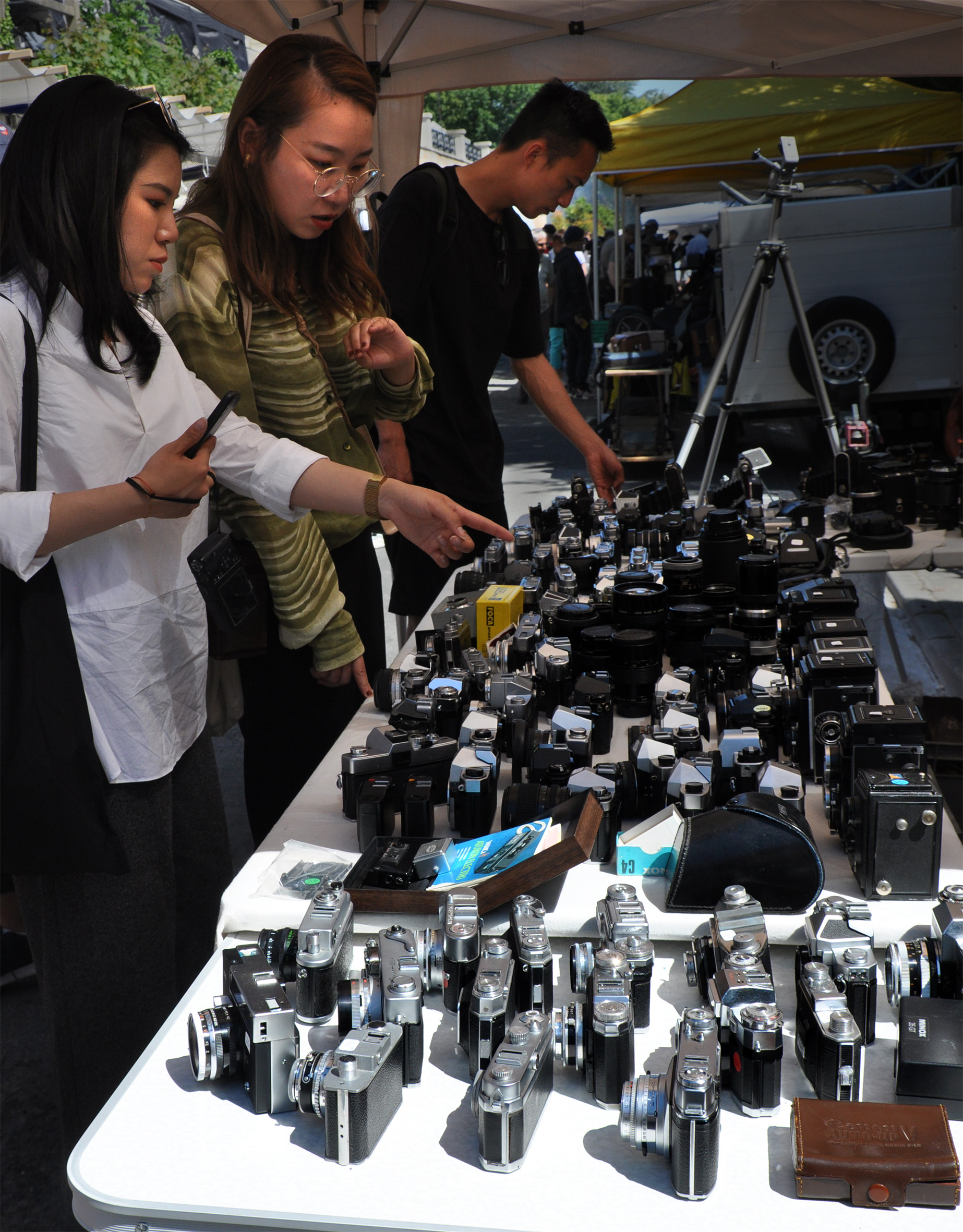 Two young women study a table densely covered in vintage 35mm film cameras, one pointing at a particular model while a third visitor browses in the background.