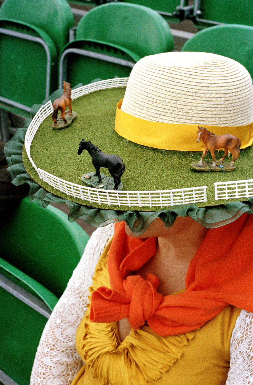 A racegoer at Ascot, seen from above, wears a wide-brimmed straw hat transformed into a miniature racecourse, complete with artificial turf, white fencing and small horse figurines.