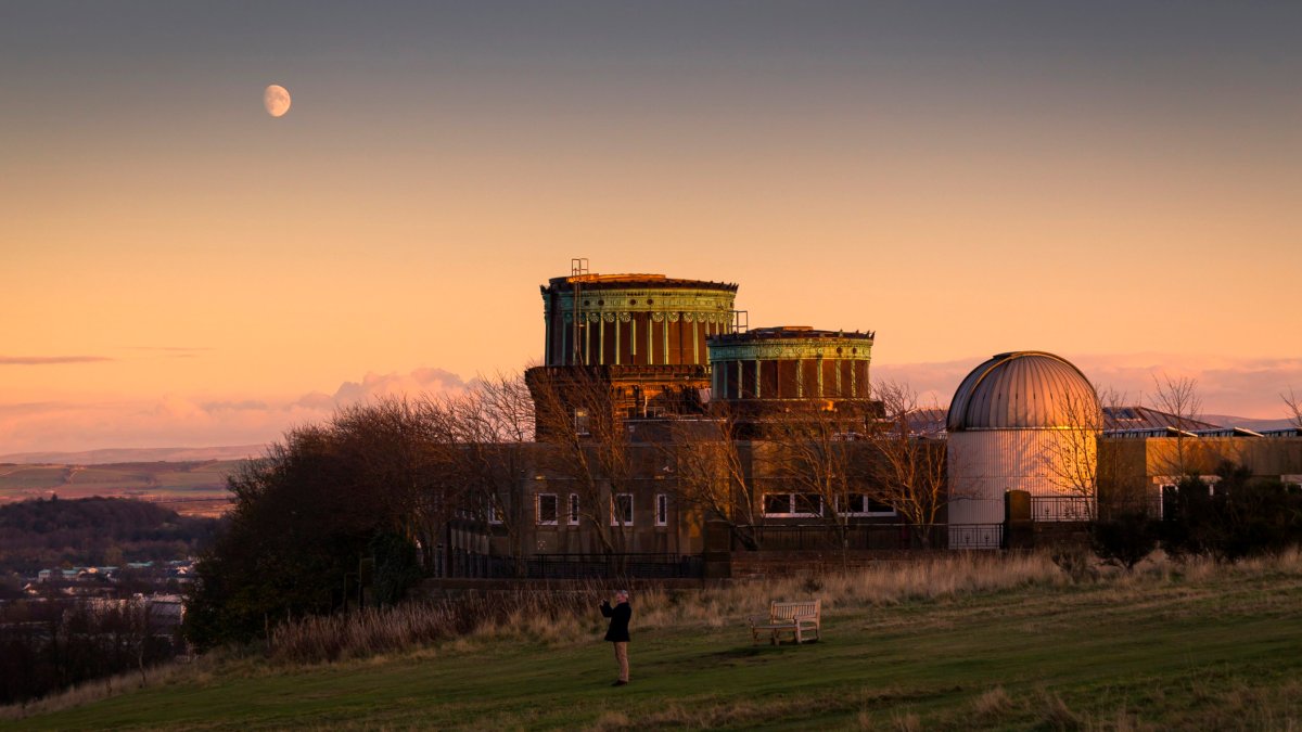 The Victorian buildings of the Royal Observatory in Braid Hill at dusk with the moon in the sky during a warm winter evening, a man taking a picture at the stunning sunset pink light over the north of Edinburgh.
