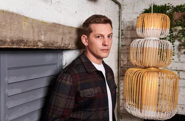 James Sutton posing as his Hollyoaks character John Paul McQueen, against a wall.