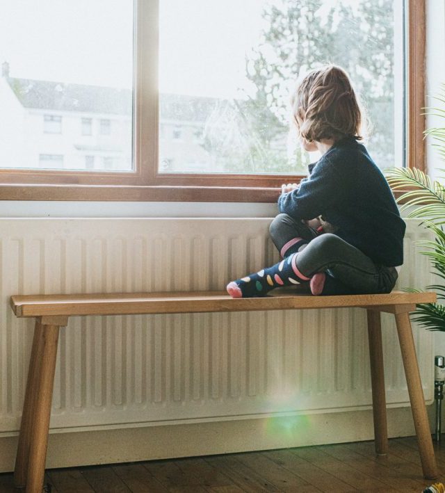 A young girl plays sits on a wooden bench beside a long radiator and a sunny window. She gazes out through the glass. The room is homely and comfortable. Conceptual with space for copy.