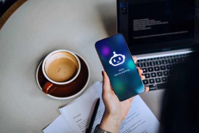 Overhead shot of businesswoman using AN AI chatbot virtual assistance app on smartphone to assist her while working on laptop. Using AI technology at work. Online robot system. Concept of VR and AR technologies. People, AI, innovation and technology