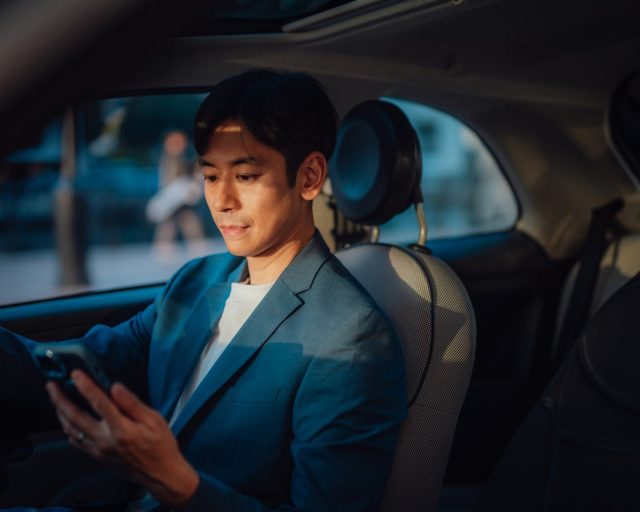 Young Asian businessman looking at mobile phone, sitting in a car. Man of style and status in full using smartphone, traveling by a car. Business travel. Mobile navigation on smartphone.
