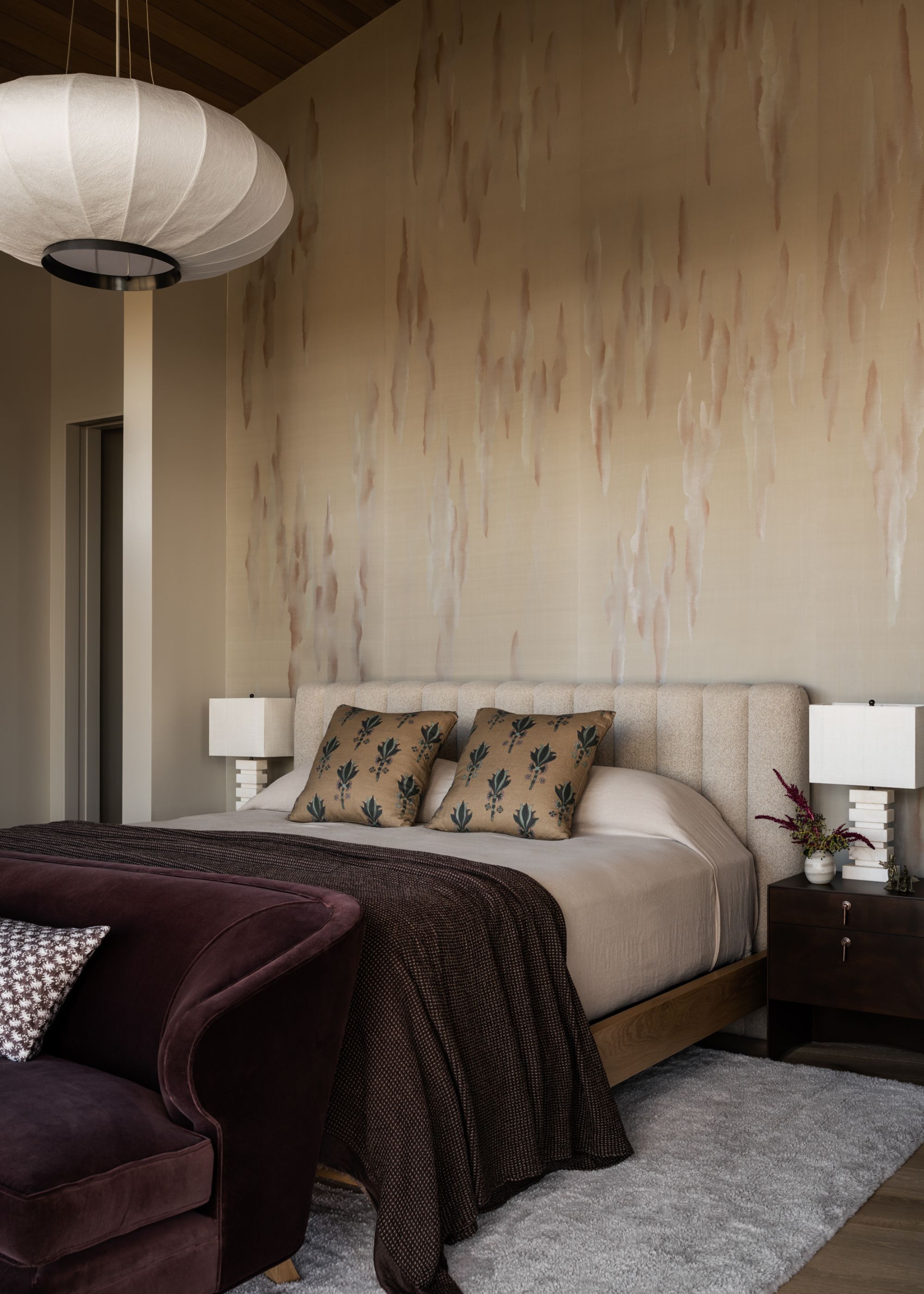 A neutral modern bedroom with a purple sofa at the foot of the bed blending with the throw at the base of the bed below two throw pillows and nightstands either side with white table lamps