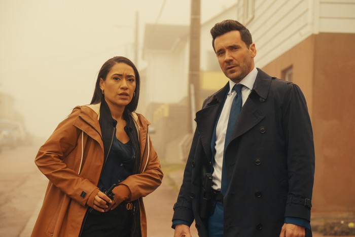 SP_EP108_D12_DC_065 Josephine Jobert and Allan Hawco in Saint Pierre, stood next to each other. She is wearing a black T-shirt and brown coat while he is wearing a black coat and a suit.