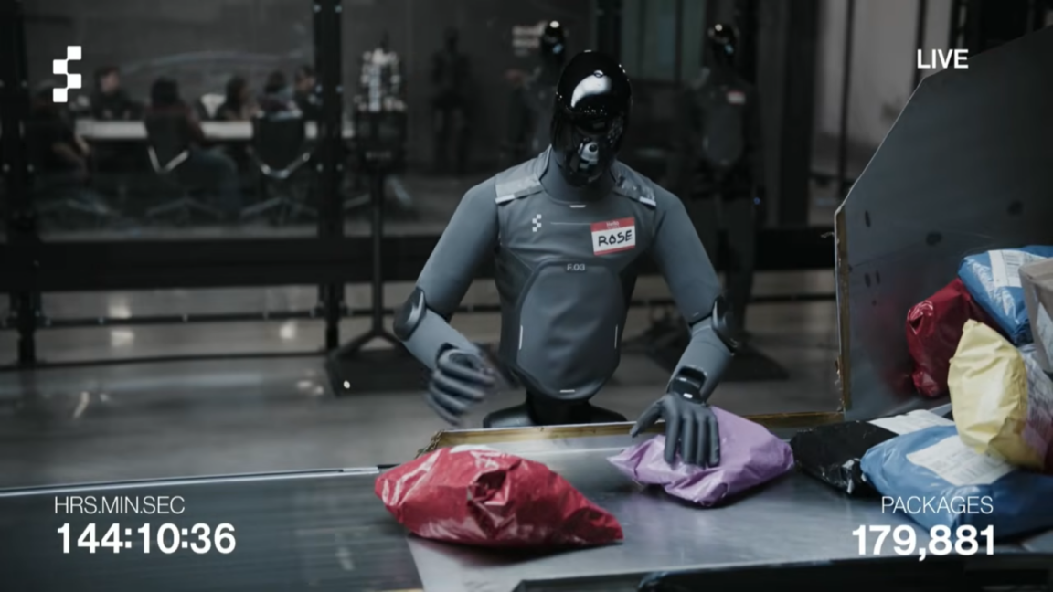 A humanoid robot with a gray body and black, reflective head stands next to a package sorting station while moving boxes and padded envelopes onto a conveyor belt.