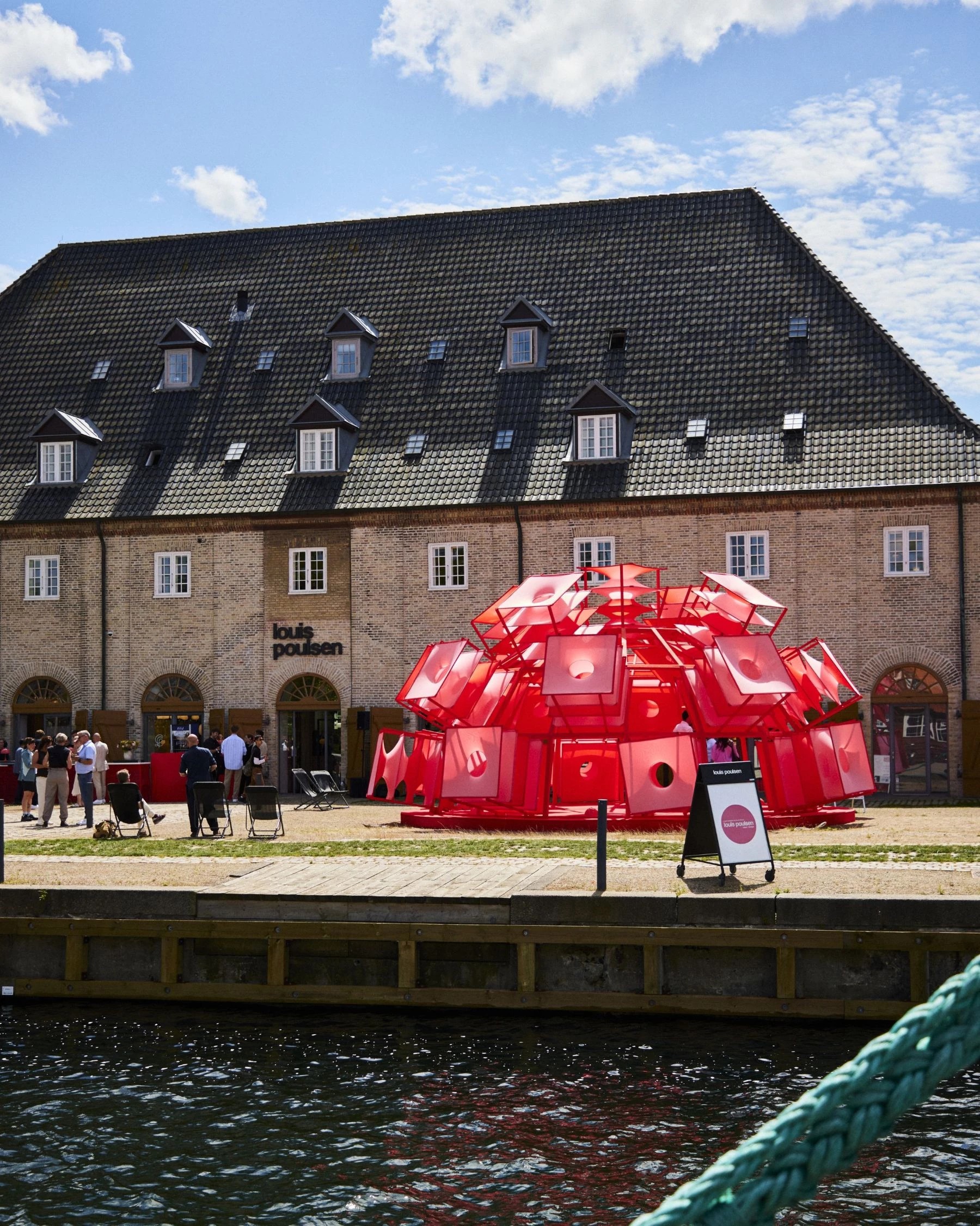 Red, multi-part outdoor installation looking like a spaceship, placed in front of an old building in brick overlooking a river in sunshine.