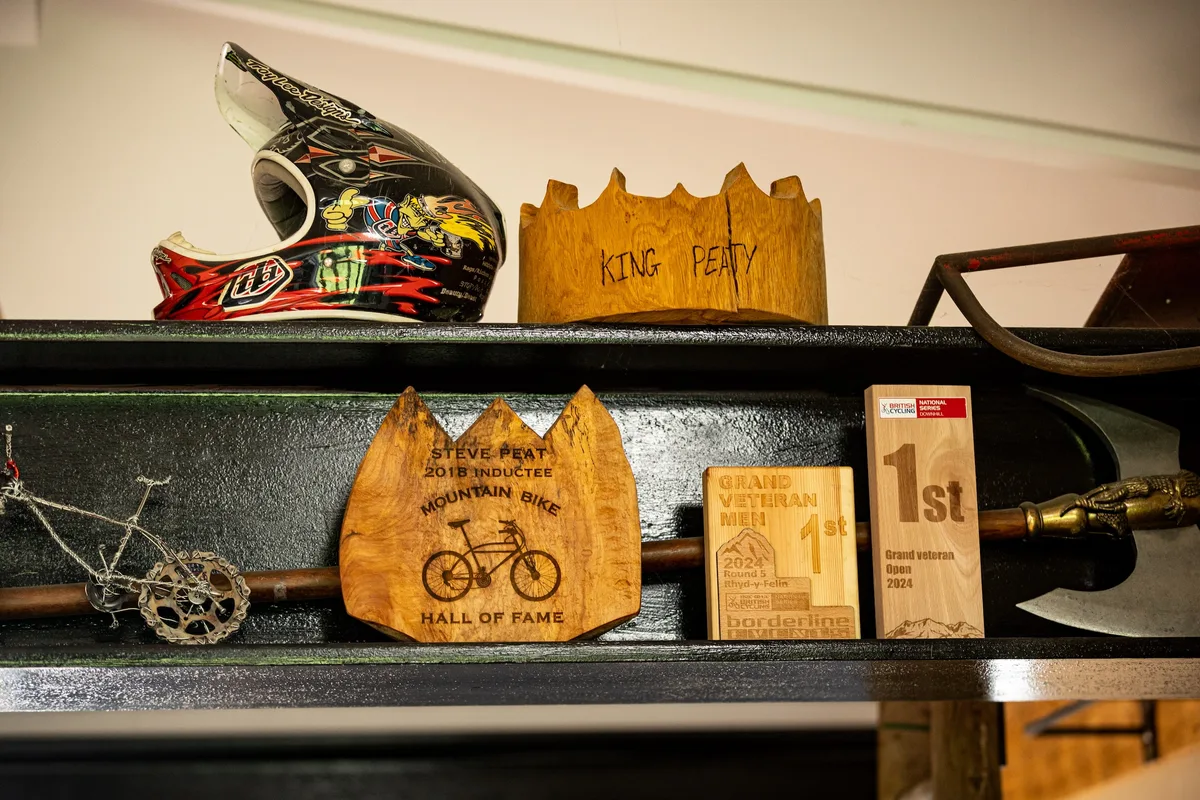 Steve Peat’s mountain bike trophies and memorabilia displayed on shelves, including a Mountain Bike Hall of Fame plaque, race awards and a Troy Lee Designs helmet.