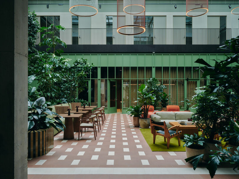 Modern indoor courtyard with green walls, potted plants, lounge chairs, tables, and circular ceiling lights arranged in a spacious seating area.