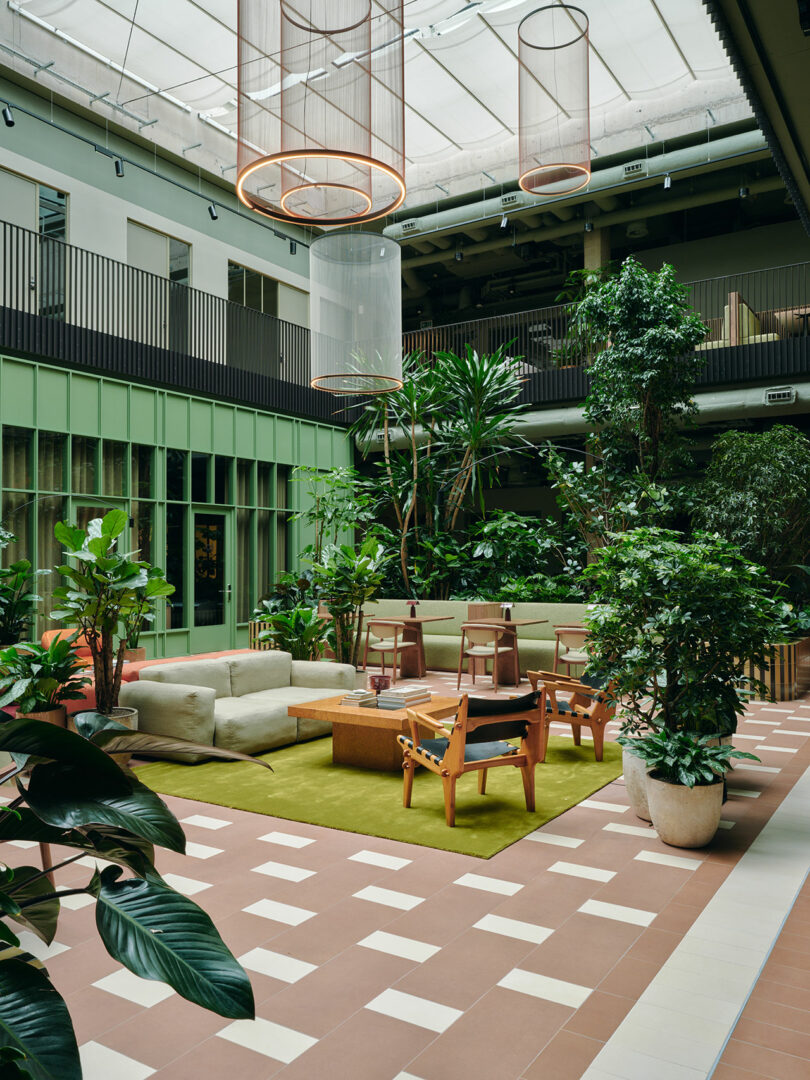 Modern indoor lounge area with green plants, tan sofas, wooden chairs, and large skylights; suspended light fixtures and tiled floor are visible.