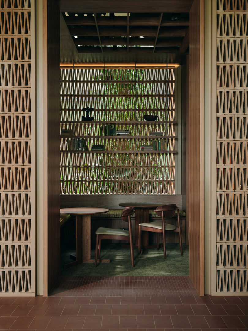 A modern room with wooden geometric lattice walls, a large window, a table, and three wooden chairs, with greenery visible outside.