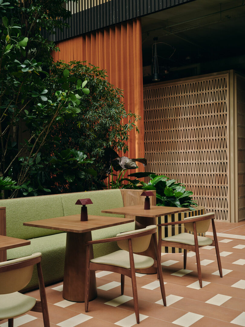 A modern café interior with wooden tables, green upholstered chairs and sofa, indoor plants, and geometric-patterned wall dividers.