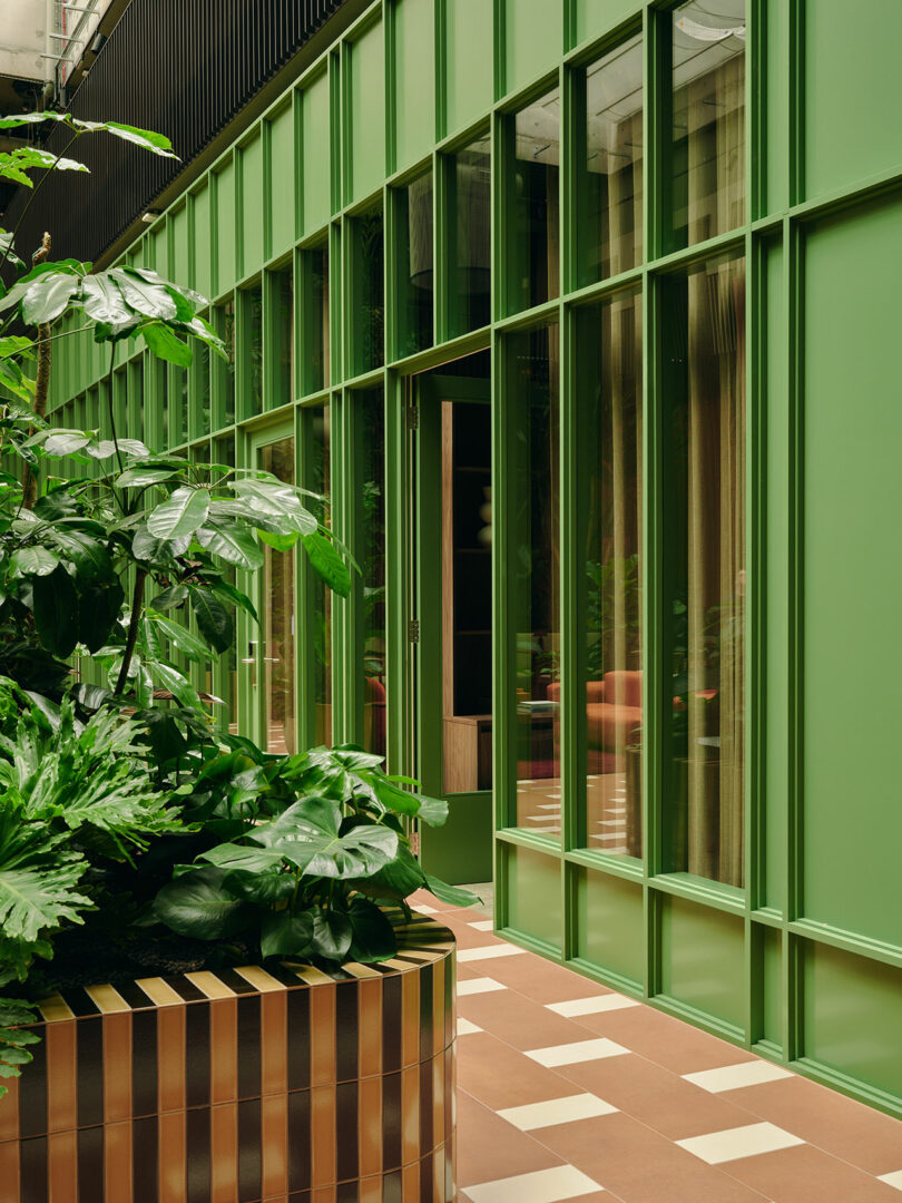 Green-framed glass doors and windows line a corridor with large leafy plants in a modern planter and a tiled brown and white floor.