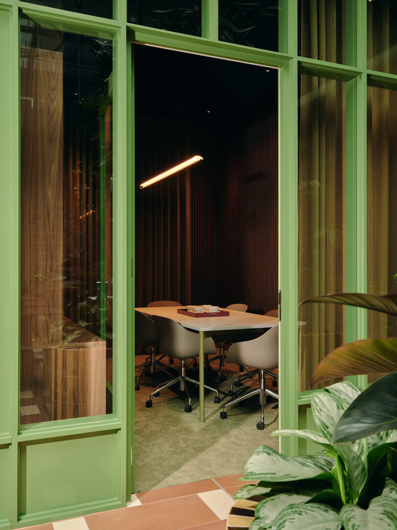 A small meeting room with a table, five chairs, a rectangular light overhead, and floor-to-ceiling green-framed glass windows. Some plants are visible in the foreground.