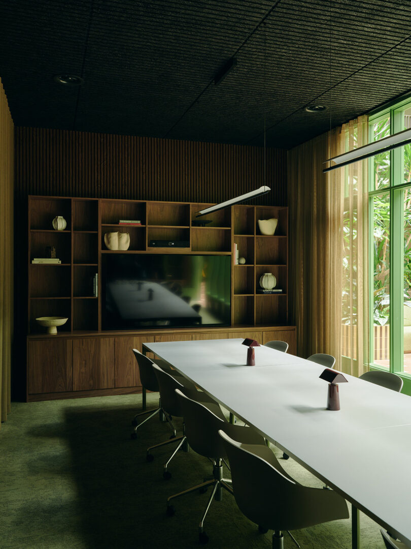 A modern conference room with a long white table, white chairs, a built-in wooden shelf with decor, a large TV, and floor-to-ceiling windows overlooking greenery.