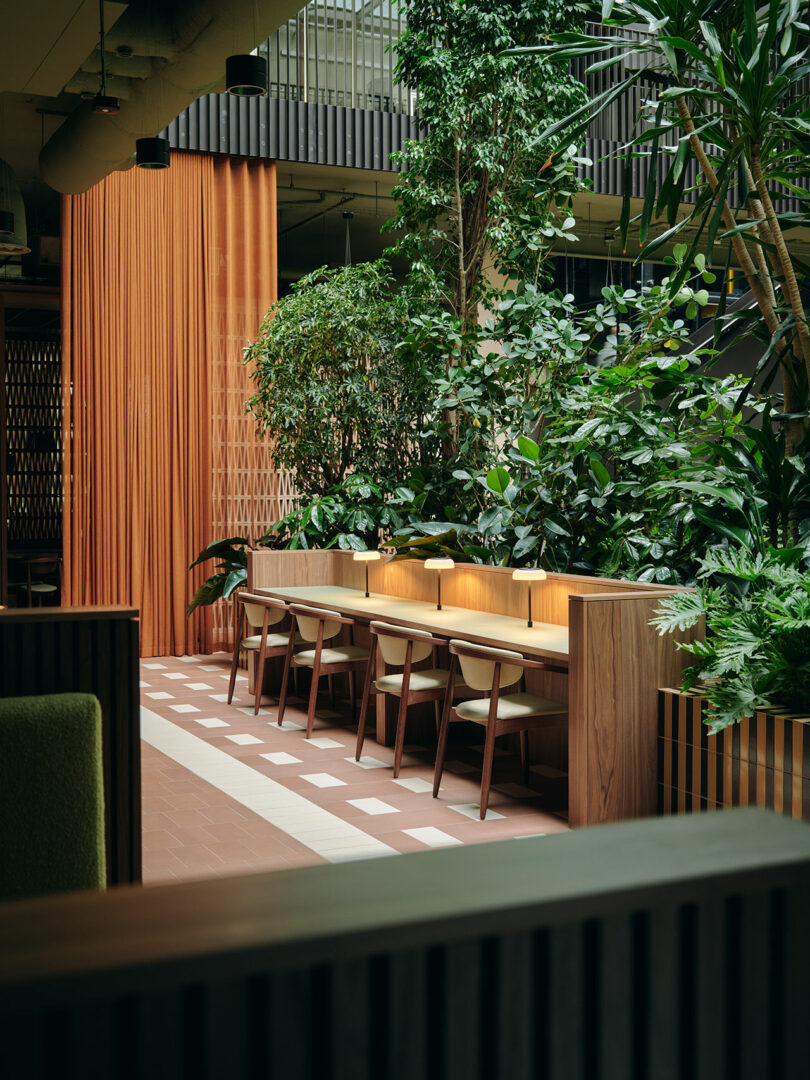 A modern indoor seating area with wooden desks, chairs, desk lamps, and abundant green plants, separated by an orange curtain and tiled flooring.