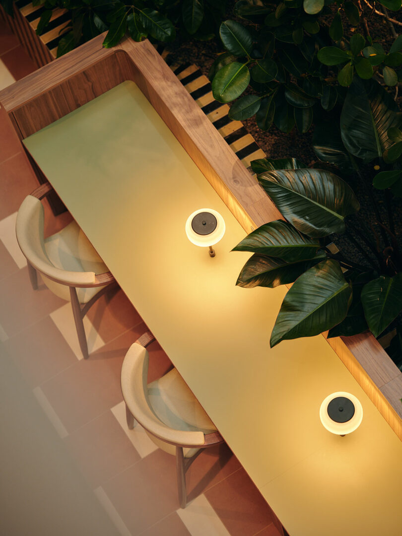Overhead view of a modern seating area with two chairs, a light yellow table, two small lamps, and large green plants nearby.