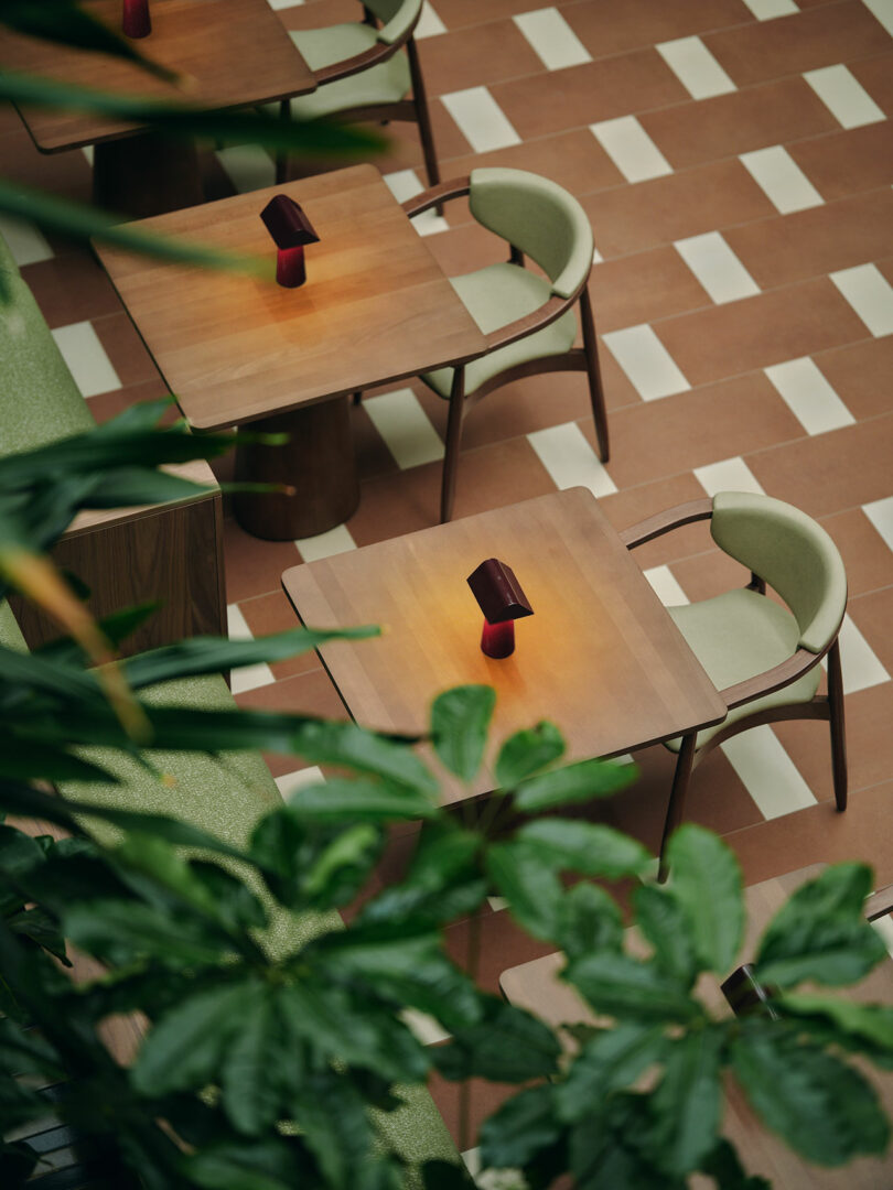 Overhead view of neatly arranged wooden tables and chairs on a patterned floor, with small red lamps on each table and green plants in the foreground.