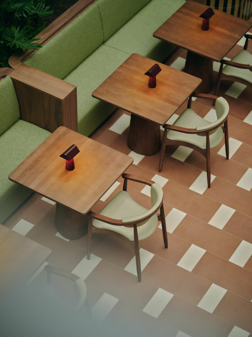 Aerial view of a modern cafe with wooden tables, green cushioned chairs, and red small lamps on each table, set on a tiled floor with alternating beige and terracotta stripes.