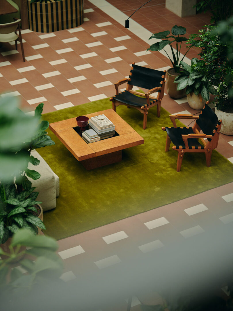 Modern sitting area with a square wooden coffee table, two black lounge chairs, books, and potted plants on a green rug over a tiled floor.