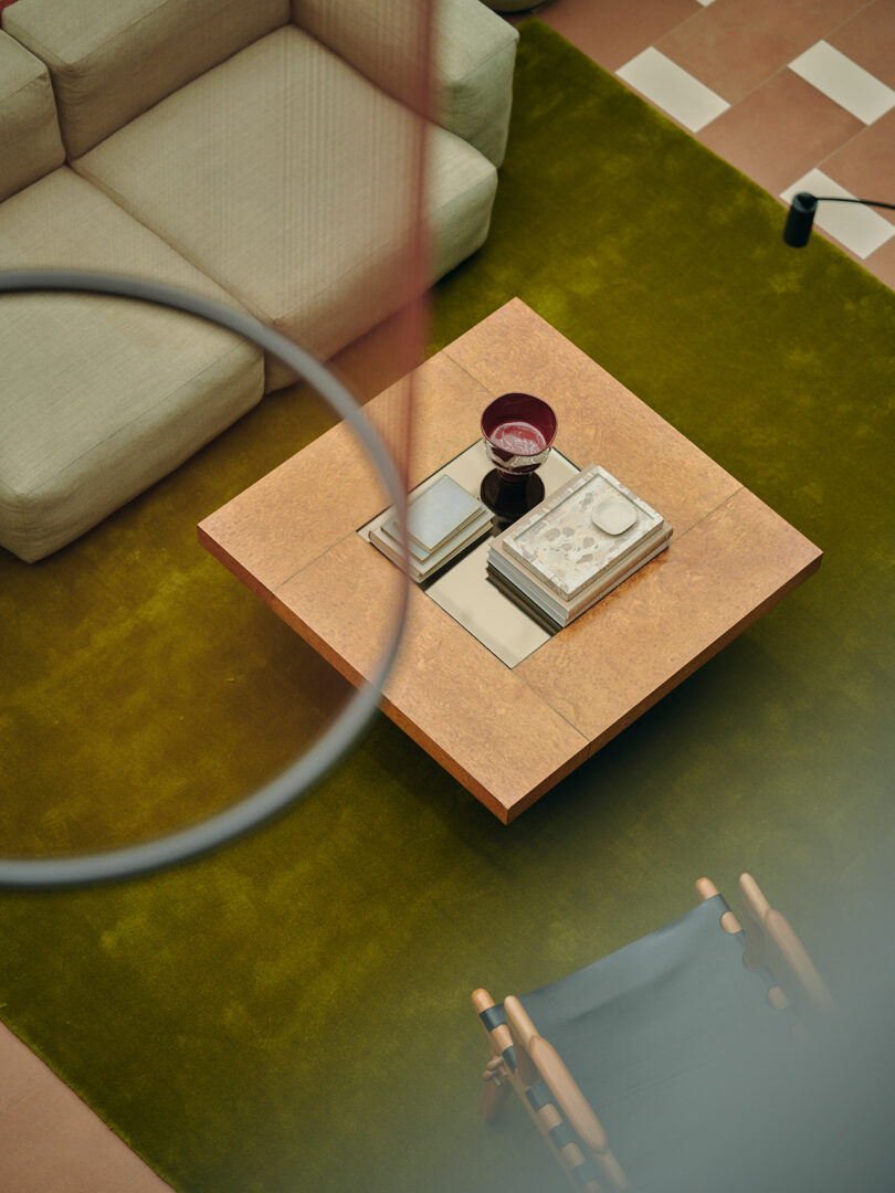 A square coffee table with books, a small dish, and a glass is set on a green rug beside a beige sofa and a wooden chair.