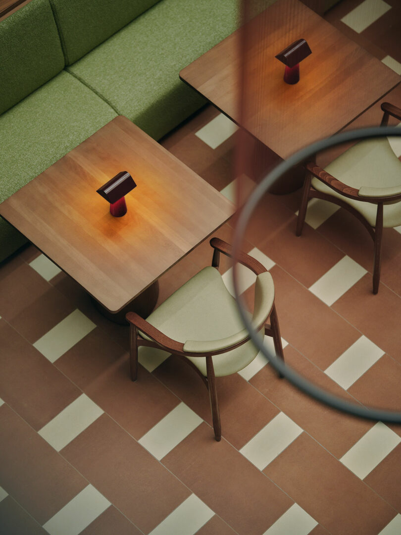 A green cushioned bench and a wooden chair sit at a wooden table with a small modern lamp, all on a tiled floor with a geometric pattern.