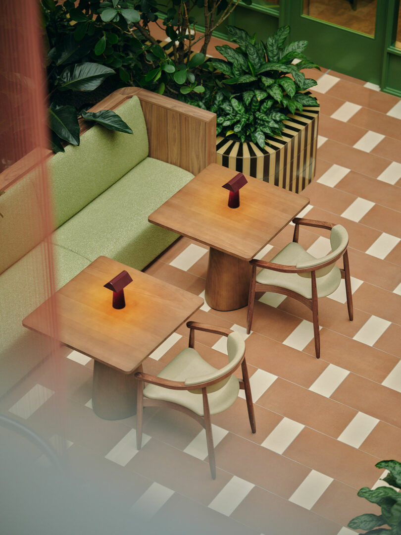 Two wooden tables with small lamps, cushioned chairs, and a green sofa are arranged in a tiled seating area surrounded by plants.