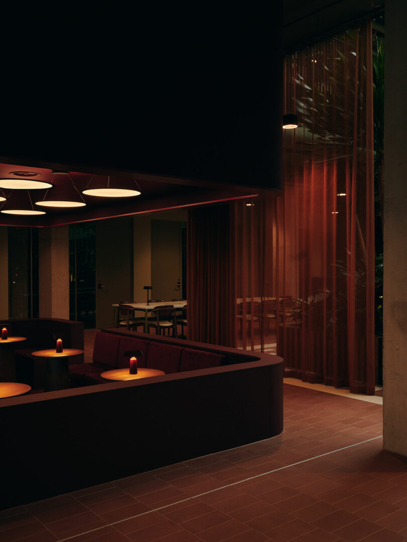 A dimly lit modern lounge with round tables, dark seating, red curtains, and pendant lights, adjacent to a dining area visible through an open doorway.
