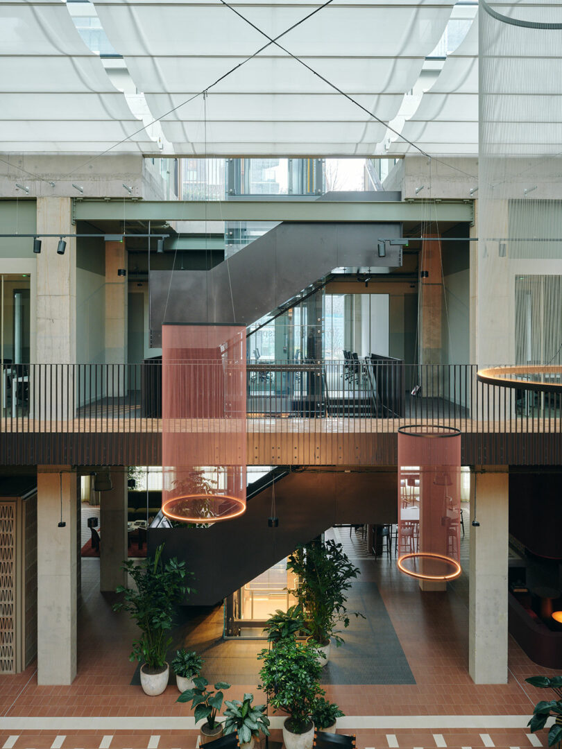 Modern indoor atrium with central black staircase, large windows, indoor plants, and circular hanging light fixtures; upper and lower levels visible.