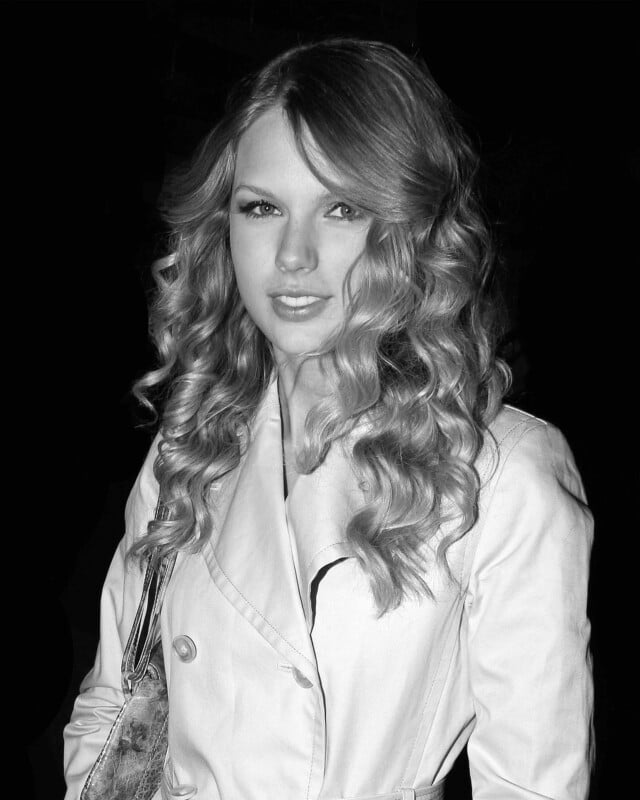 A woman with long, curly hair wearing a light-colored trench coat and carrying a shoulder bag, posing and looking at the camera against a dark background.