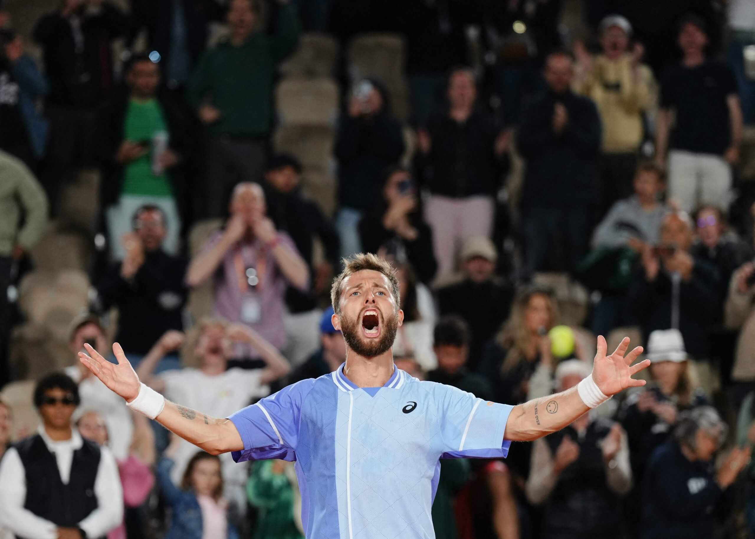 Corentin Moutet roars in delight wearing a blue tennis outfit with a crowd behind him.