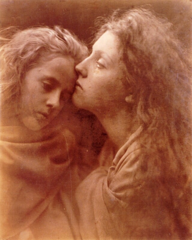 Two young people with long hair, wrapped in soft fabric, sit closely together. One rests their head on the other's shoulder while the other gently leans in, creating an intimate and tender moment. The image is softly toned and vintage.