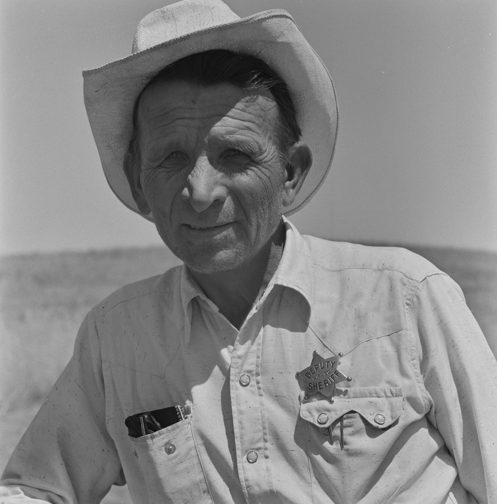 A weathered older man wearing a cowboy hat and a deputy sheriff's star badge poses outdoors against a flat horizon.