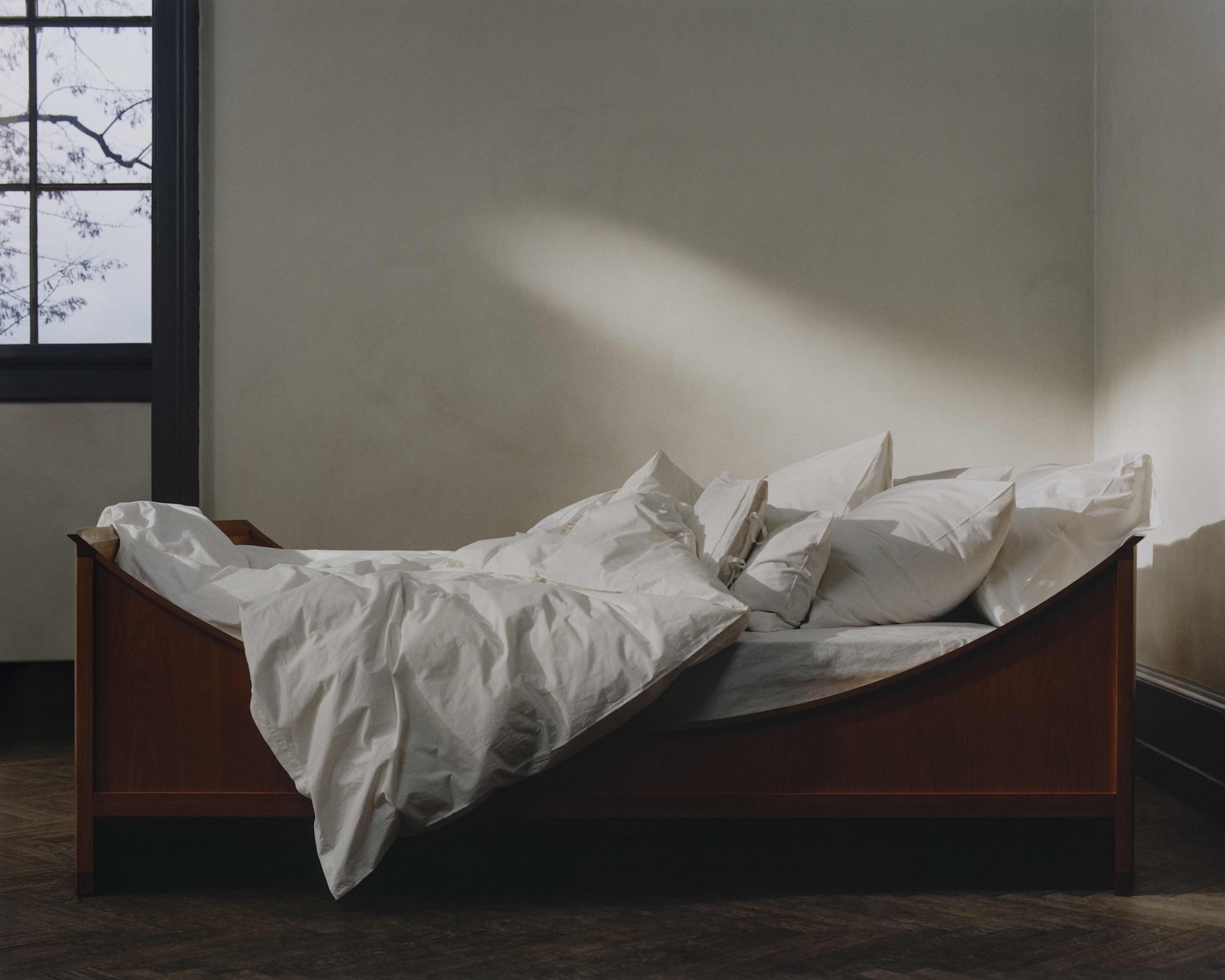 A bed with white bed sheets and a structural, chunky wooden frame.