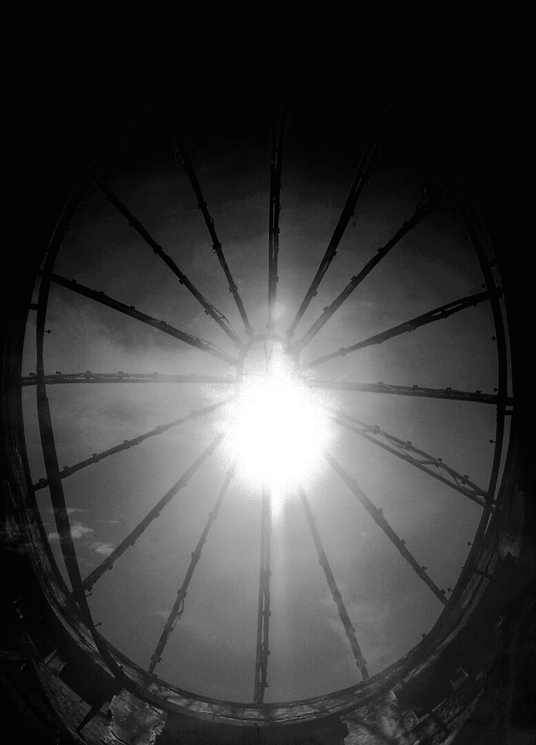 A high-contrast black and white photograph looking directly up at a bright sun through the dark, skeletal ribs of a circular architectural dome.