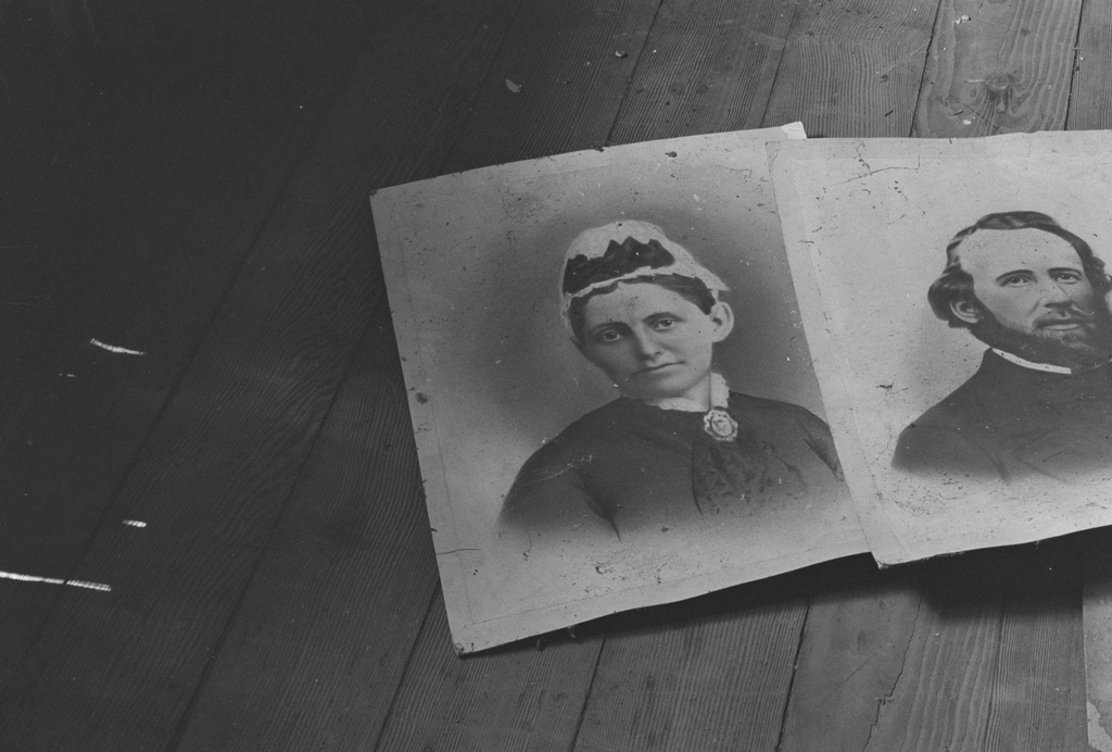 Two aged Victorian-era portrait photographs, one of a woman in a bonnet and one of a bearded man, lie side by side on a bare wooden floor.