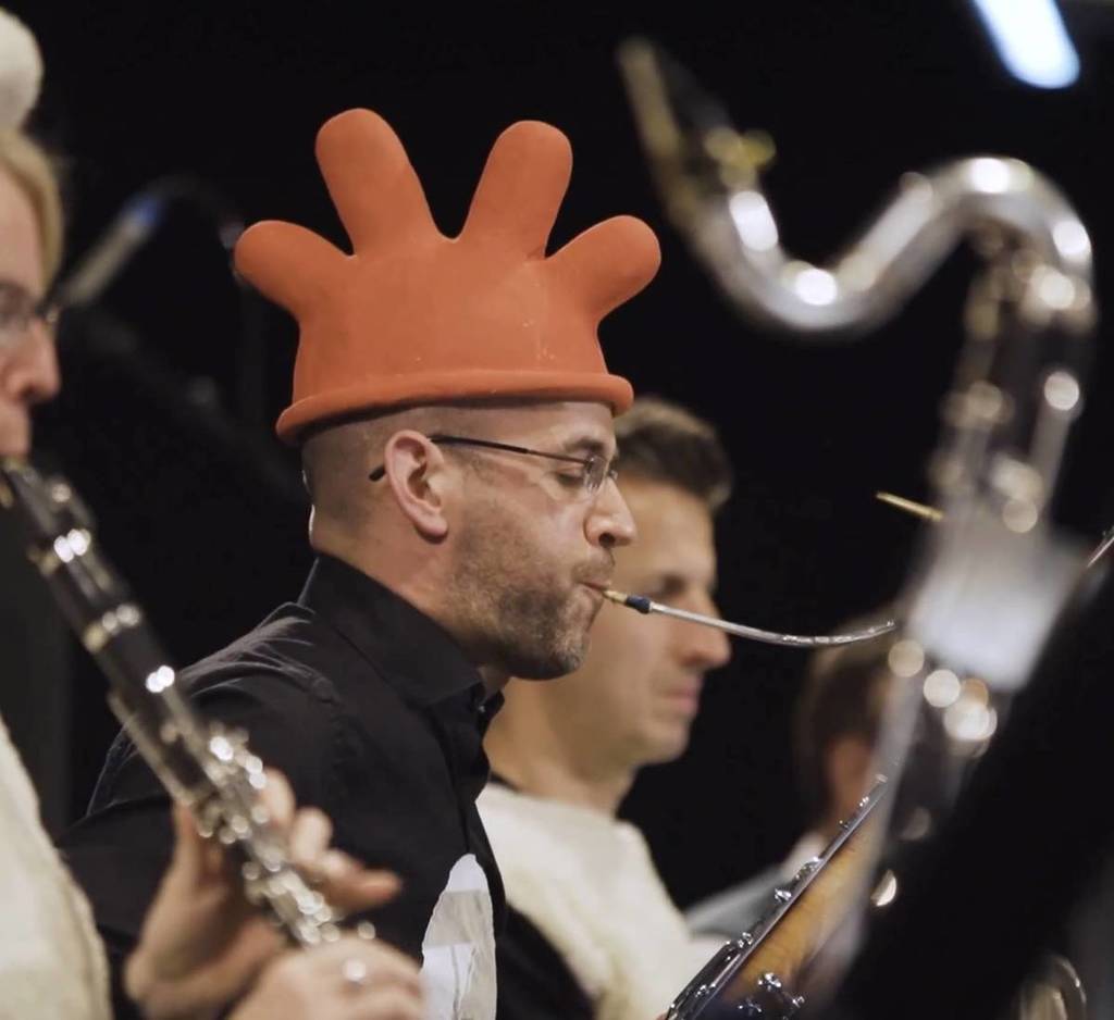 a brass instrument player playing in an orchestra while wearing a glove-type hat, similar to Feathers McGraw