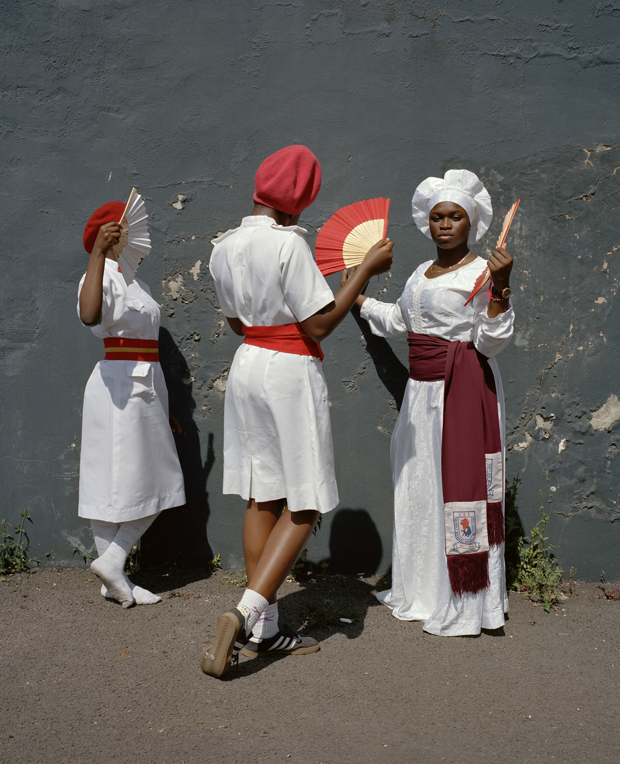 Three people in white ceremonial dress and red accessories hold folded fans against a peeling grey wall in London; one looks directly at the camera while the other two face each other in profile.