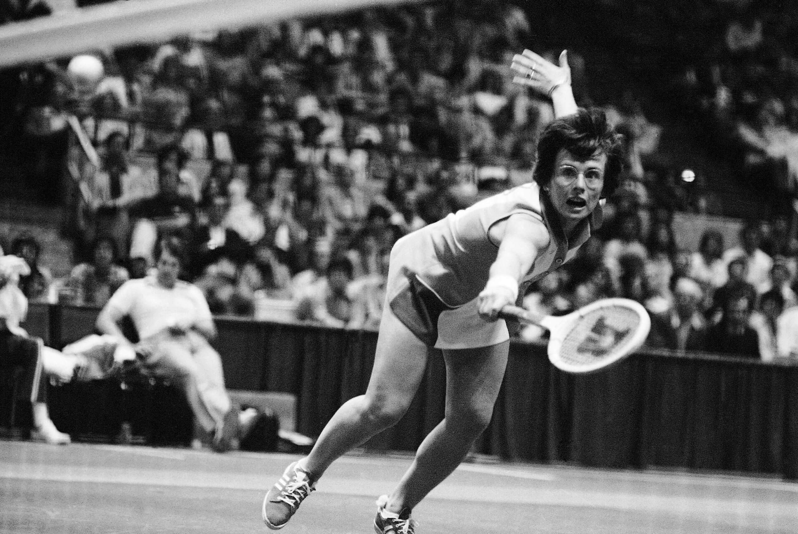 Billie Jean King stretches to hit a backhand with her right hand in a black-andwhite-photo of a tennis match with a sizeable crowd in the stands.