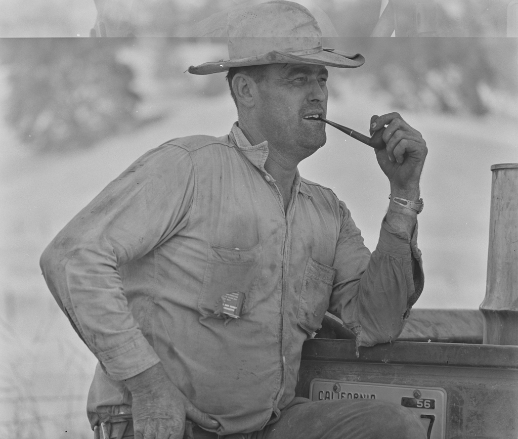 A broad-shouldered man in a dusty work shirt and cowboy hat smokes a pipe while leaning against a vehicle with a California licence plate.