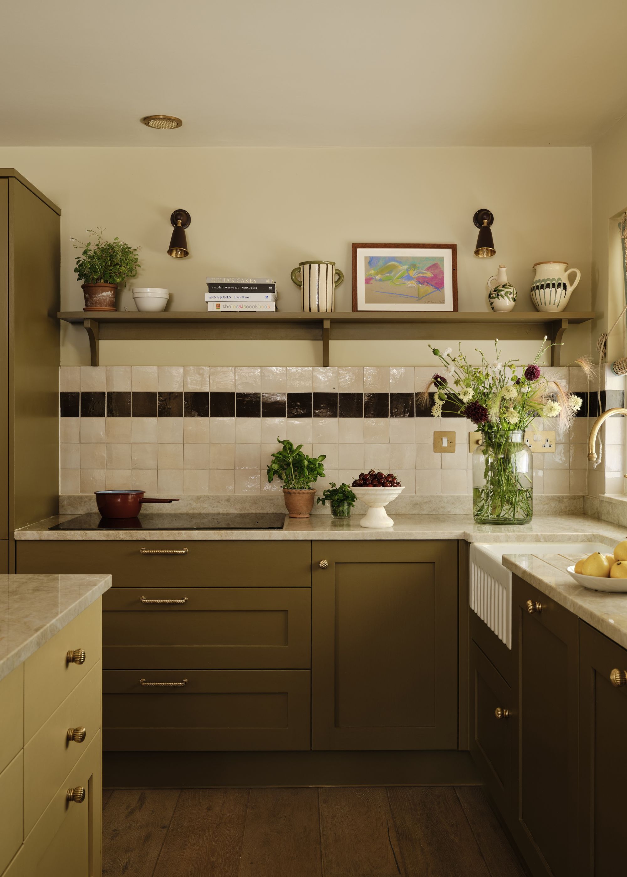 A cozy, modern kitchen with rich olive green cabinets, white Zellige wall tiles with a black accent row, white countertops and warm neutral walls with a shelf with decor.