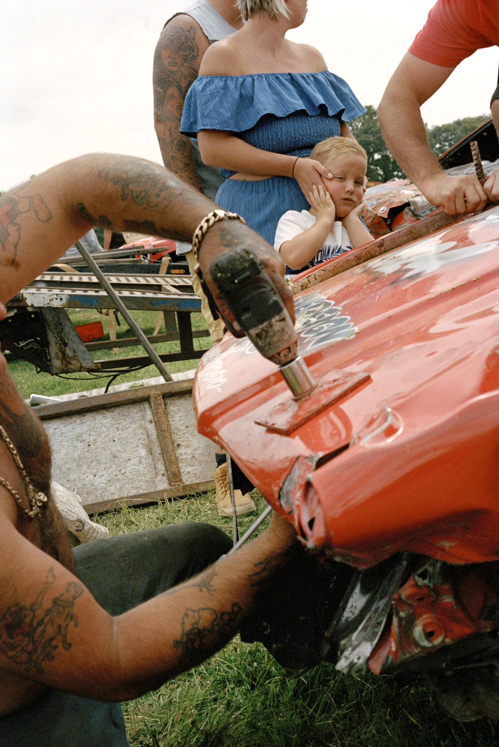 A tattooed mechanic drills into the crumpled bonnet of a red banger car while a young blonde boy watches intently, cradled by a woman in a blue off-shoulder top at Smallfields Raceway.