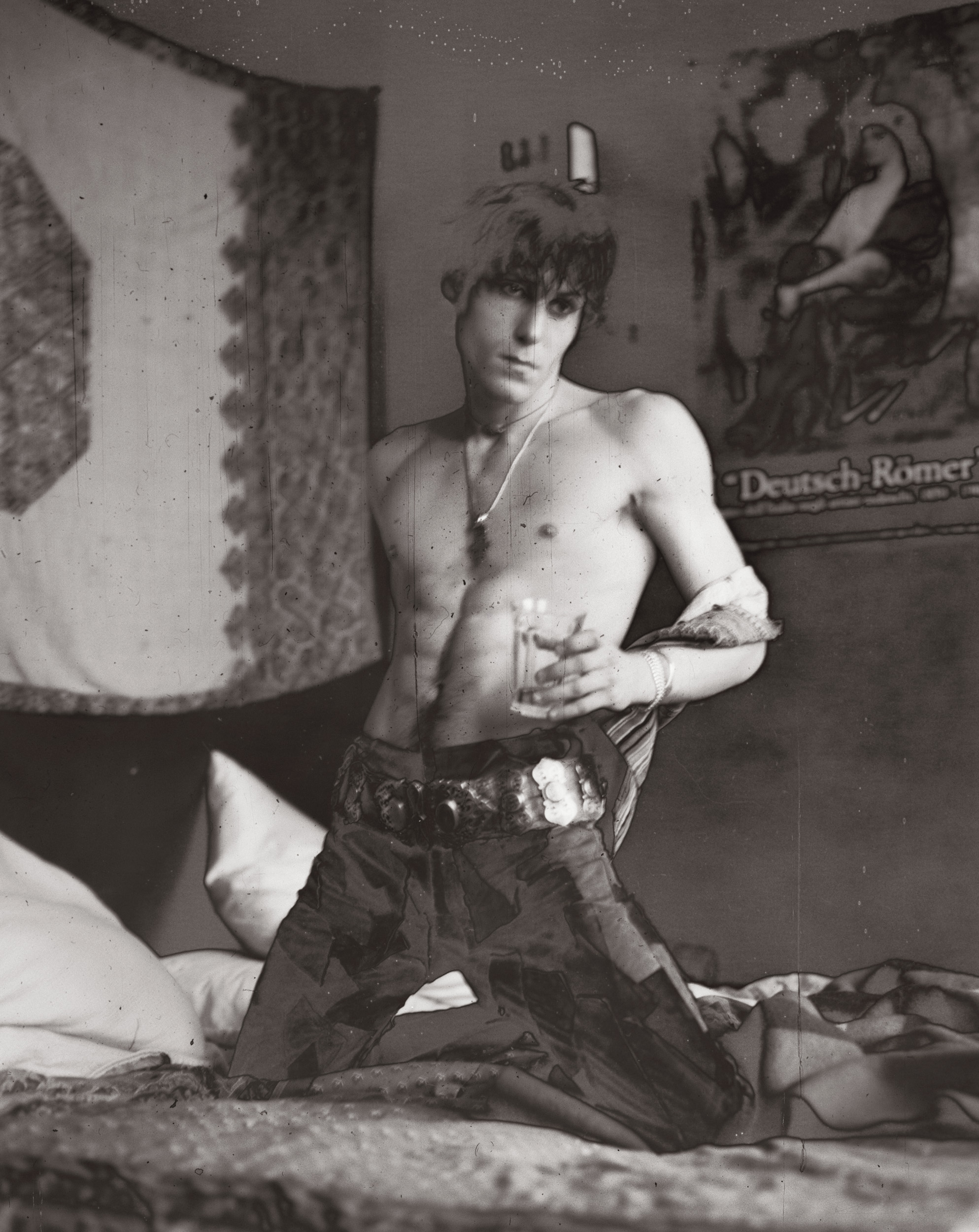 A black and white, slightly grainy photograph of a shirtless young man with tousled hair kneeling on a bed, holding a glass and wearing patterned trousers with a large decorative belt.