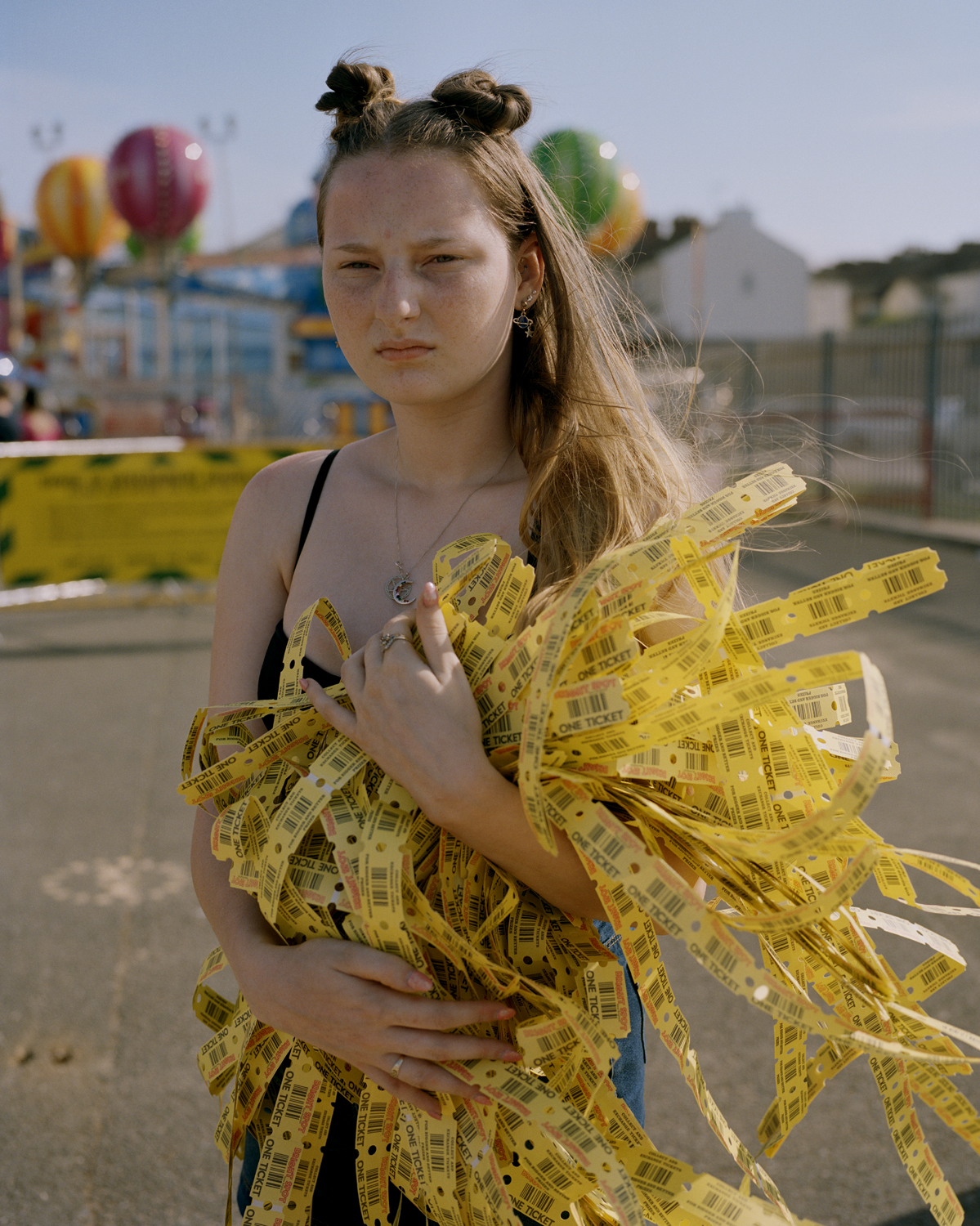 A teenage girl with double bun hair stares directly into the camera while clutching an enormous rustling bundle of yellow carnival ride tickets at New Brighton Beach fairground.