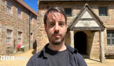 Ben Spink stands centered in a sunlit stone courtyard, facing the camera, wearing a dark shirt and jacket. Behind him are rustic buildings made of warm-toned stone, including one with pink-trimmed windows on the left and another with a thatched roof and a small wooden porch on the right. Bright blue sky and a pot of red and white flowers add colour to the scene.